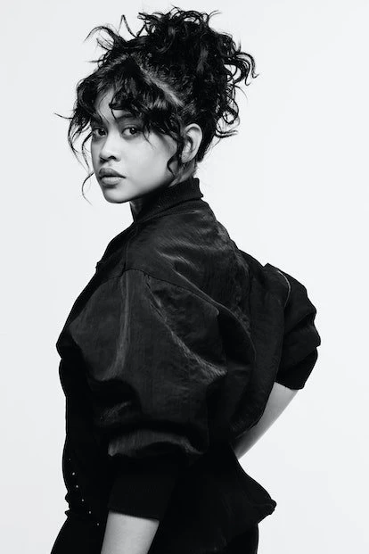 Close-up black and white portrait of a woman with curly hair, wearing a jacket, looking over her shoulder in a light studio.