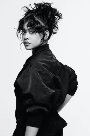 Close-up black and white portrait of a woman with curly hair, wearing a jacket, looking over her shoulder in a light studio.