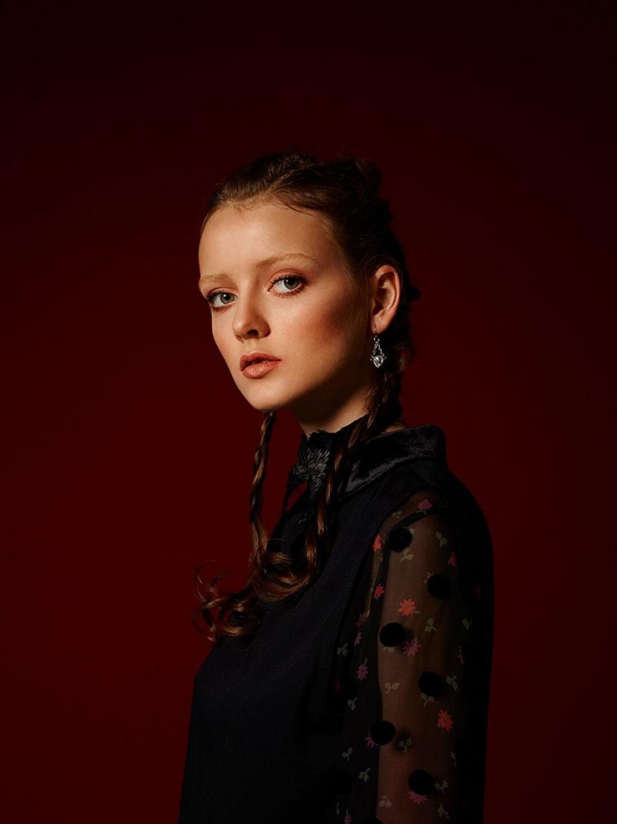 Portrait of a young woman with brown hair styled in braids, wearing a black vintage dress with floral patterned sleeves. The background is dark red.