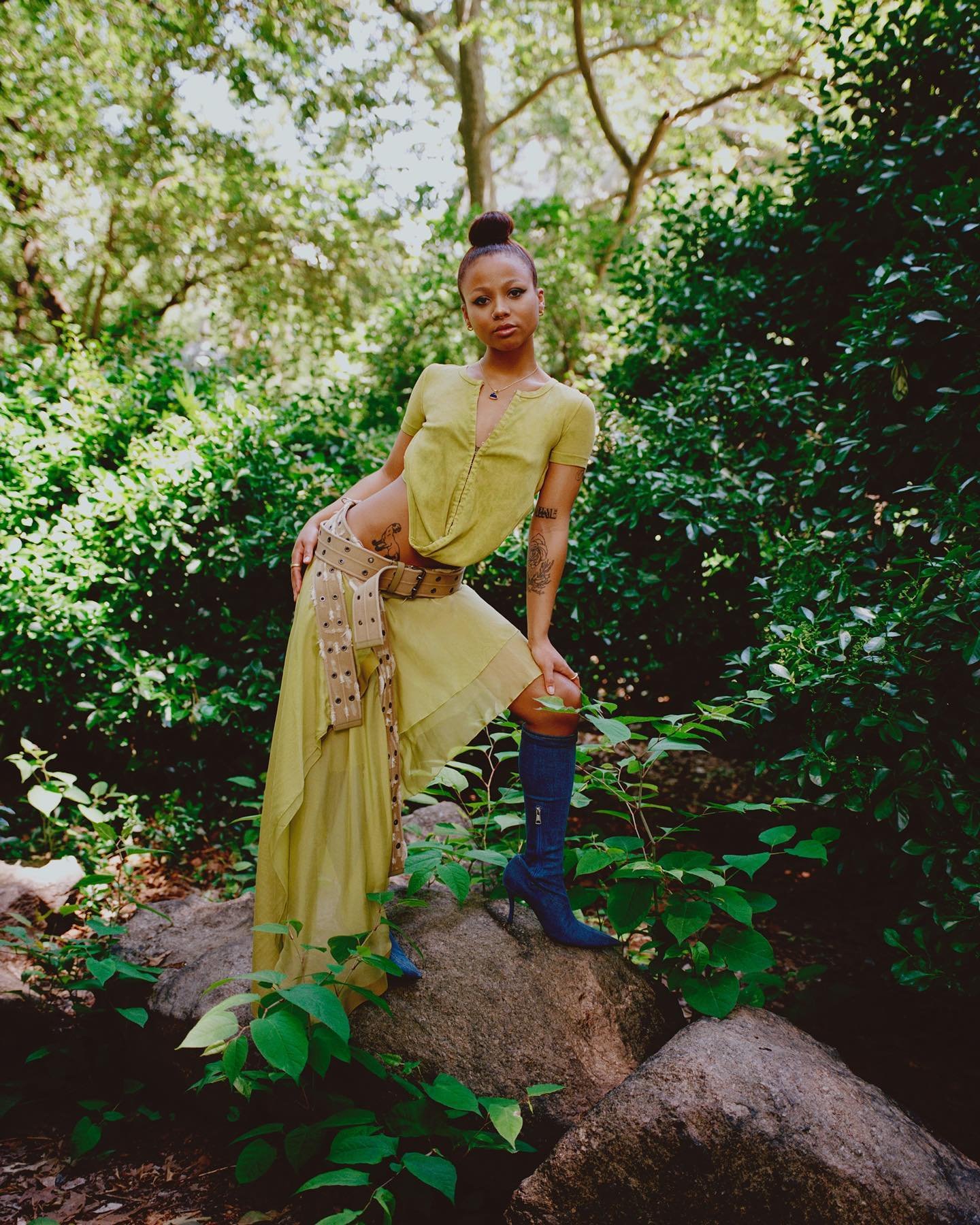 A woman with tattoos stands on a rock in a leafy forest. She is wearing a yellow outfit with blue heeled boots and a wide decorative belt.