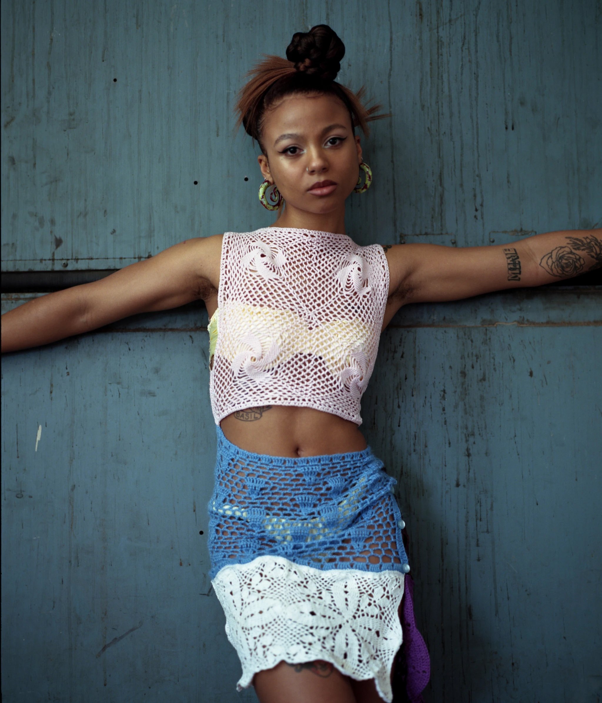 Woman in a knitted top and skirt poses against a blue textured wall. She has her arms outstretched and wears large hoop earrings.