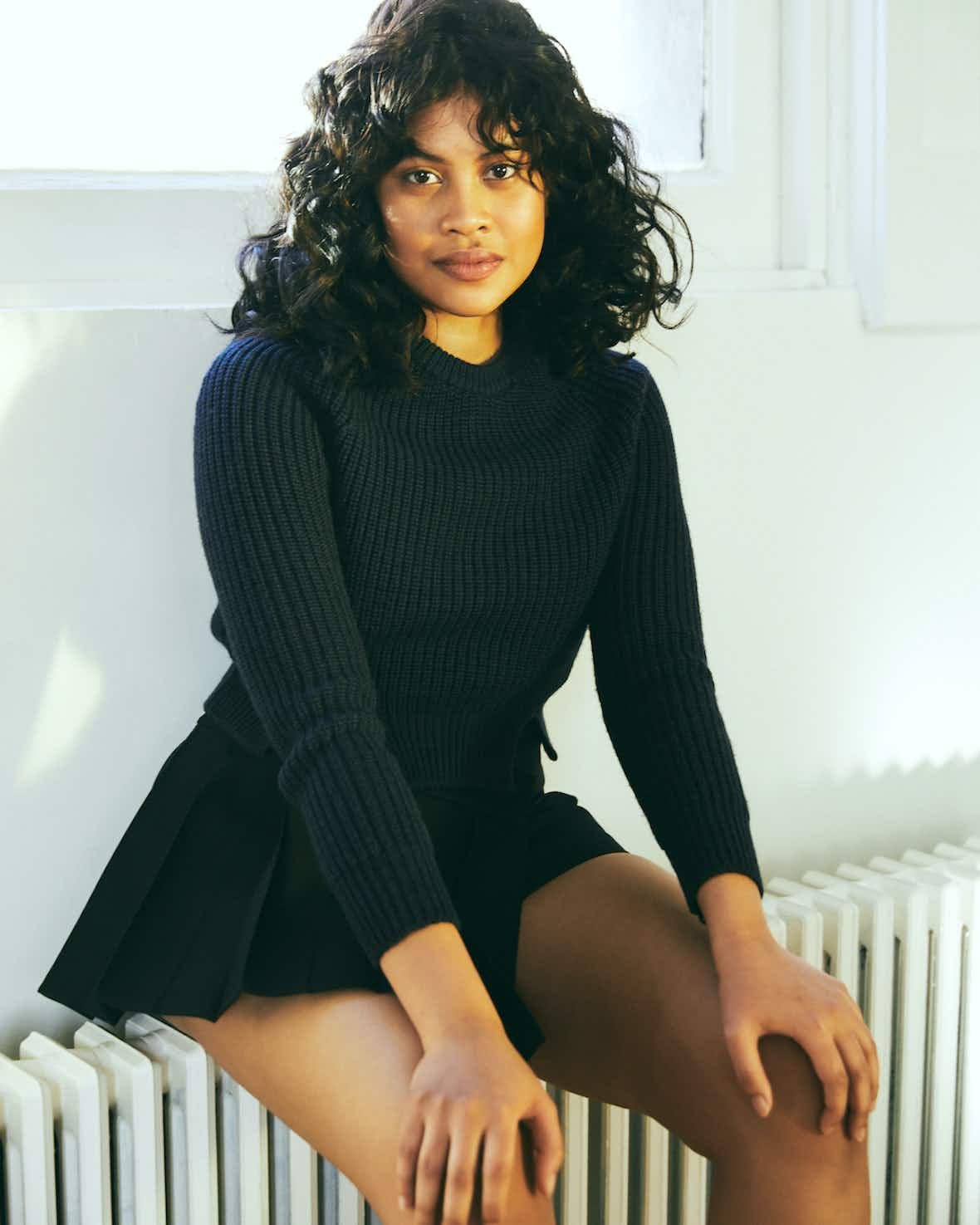 Portrait of a woman with curly dark hair wearing a knitted top and a pleated skirt, sitting indoors in front of a window.