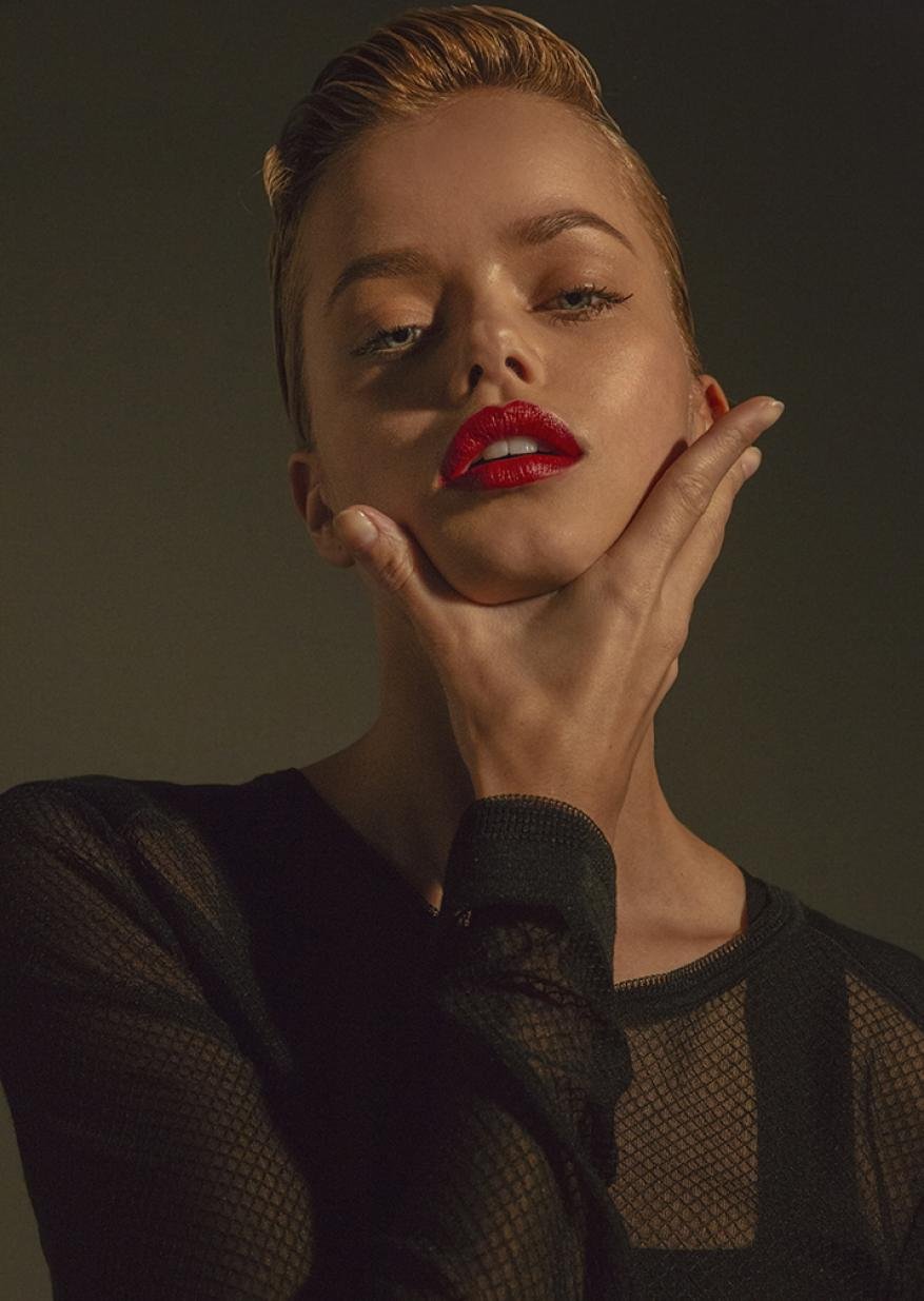 Portrait of a woman with red lipstick holding her chin with one hand, wearing a black mesh top against a dark background.