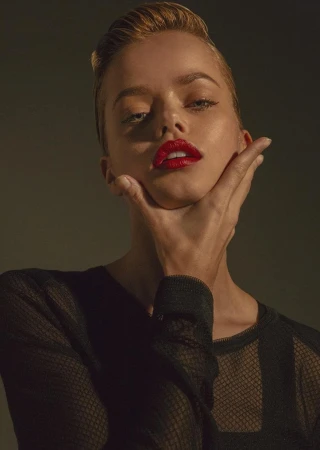 Portrait of a woman with red lipstick holding her chin with one hand, wearing a black mesh top against a dark background.