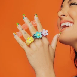 Close-up of a smiling woman displaying colourful flower rings on her fingers and vibrant nail art against an orange background