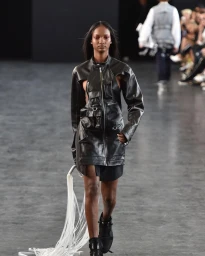 Model walking the runway wearing a black leather utility jacket and carrying white fringe accents.
