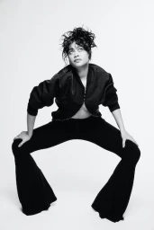 Black and white studio portrait of a woman crouching in black flared trousers and a jacket against a plain background