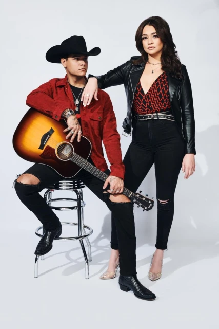 Music duo poses in studio. A man sits on a stool holding an acoustic guitar, a woman stands next to him with her hand on his shoulder.