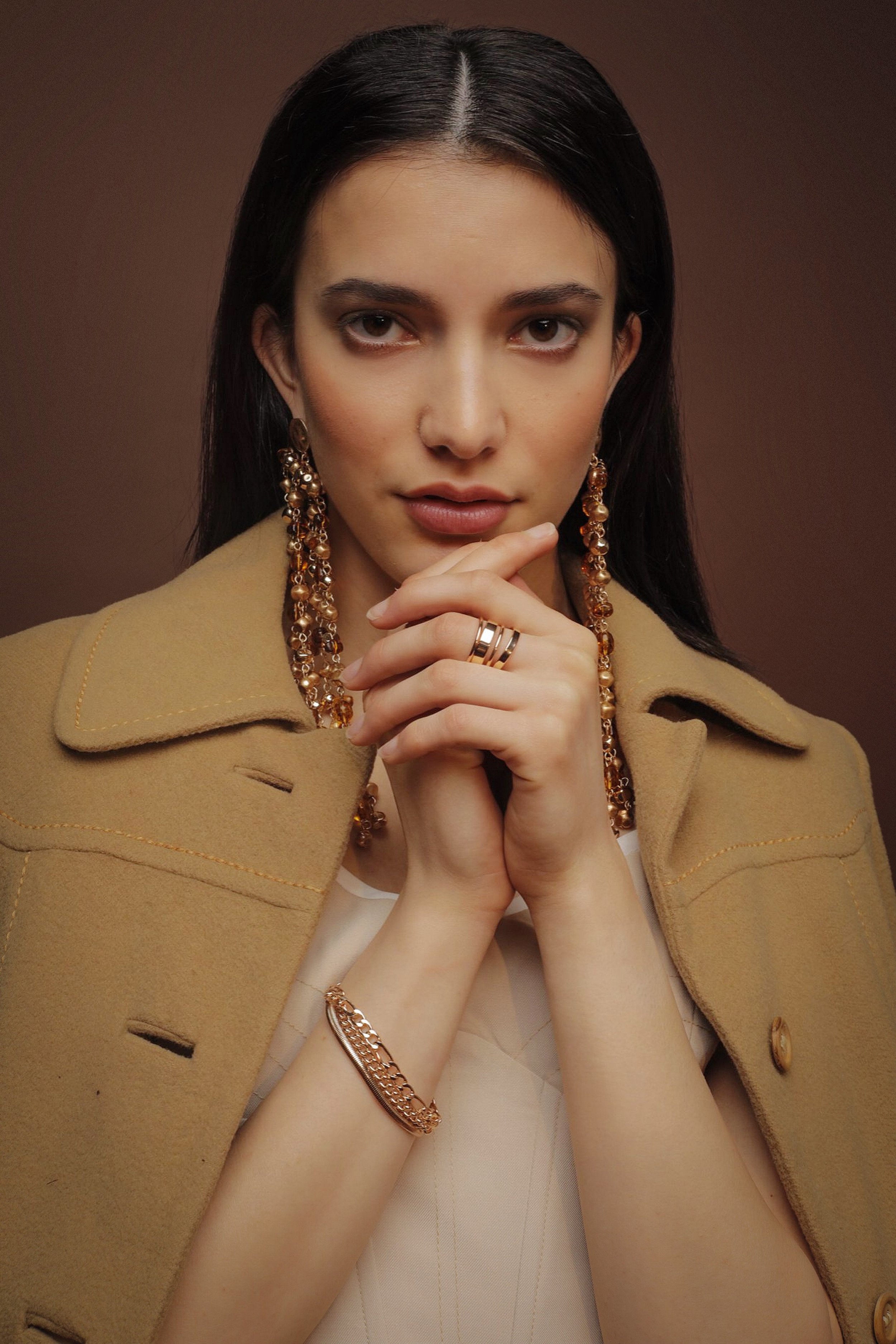 Portrait of a woman with dark hair wearing gold jewelry and a tan coat, posing against a brown backdrop in a studio setting.