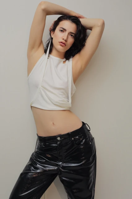 Fashion shot of a female model in a white halter top and black patent leather pants posing against a neutral wall in studio lighting.