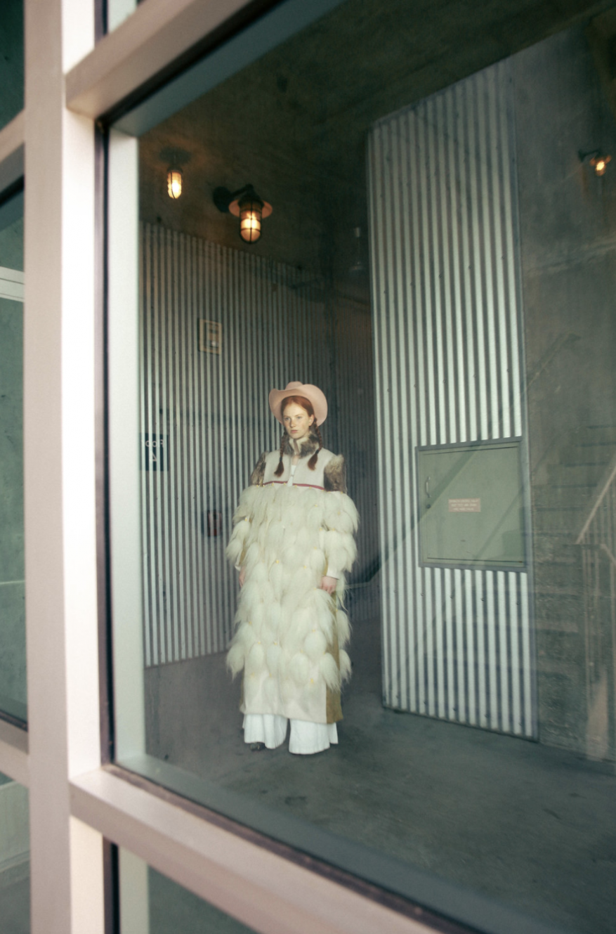 A woman in an oversized, feathery white coat and hat stands in a striped room, seen through a window. The lighting is soft and diffused.