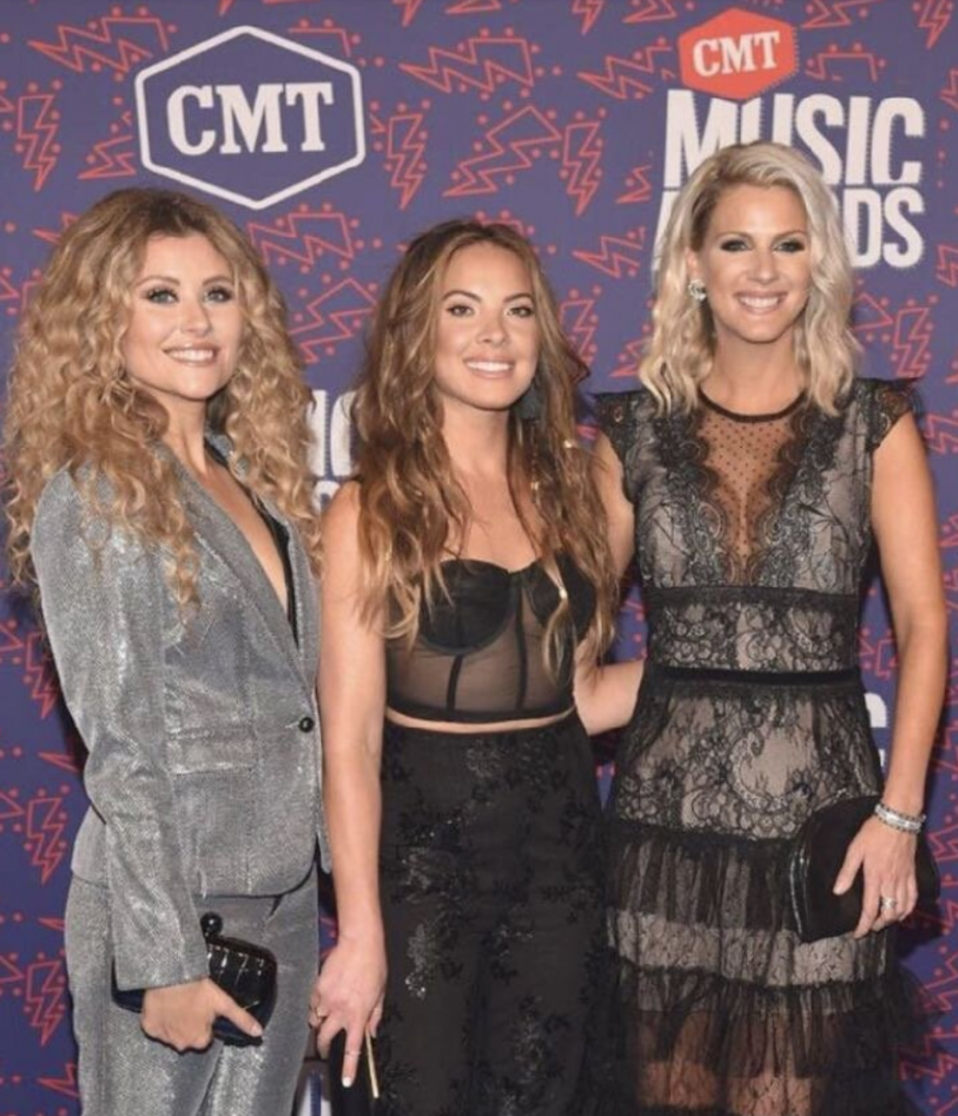 Three women pose on the red carpet at the CMT Music Awards wearing elegant evening wear against a branded backdrop.