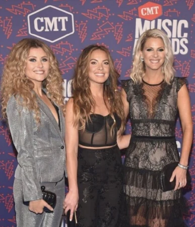 Three women pose on the red carpet at the CMT Music Awards wearing elegant evening wear against a branded backdrop.
