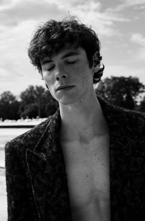 Black and white portrait of a young man with curly hair and closed eyes wearing a textured blazer outdoors.