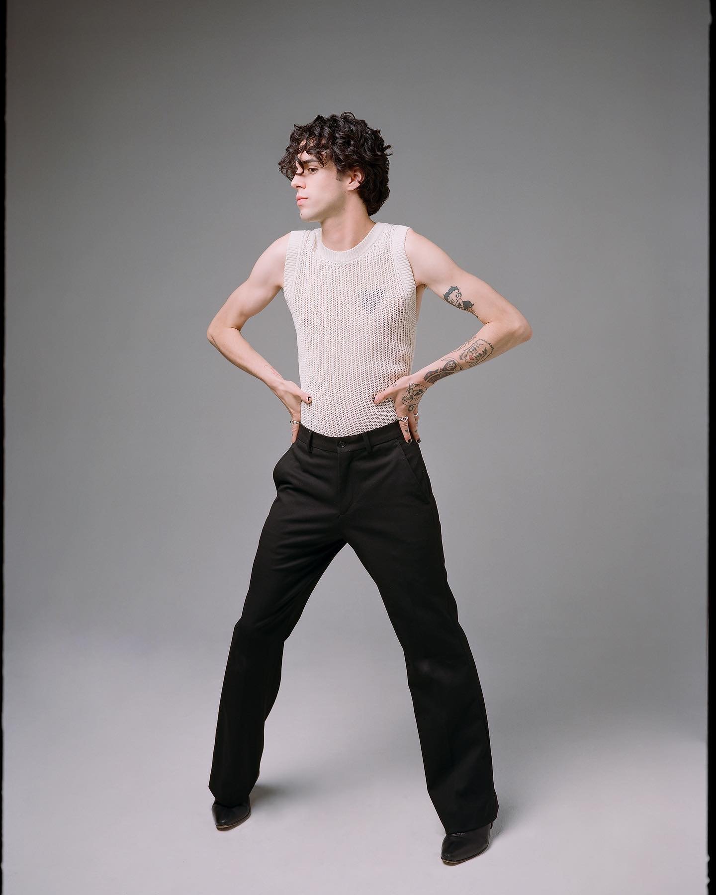 Man in textured tank top and black pants strikes a confident pose in a studio setting with a neutral background, showcasing fashion and style.