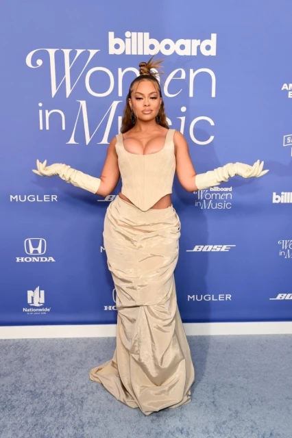 A woman in a beige corset top and skirt poses on a carpet in front of a Billboard Women in Music branded backdrop.