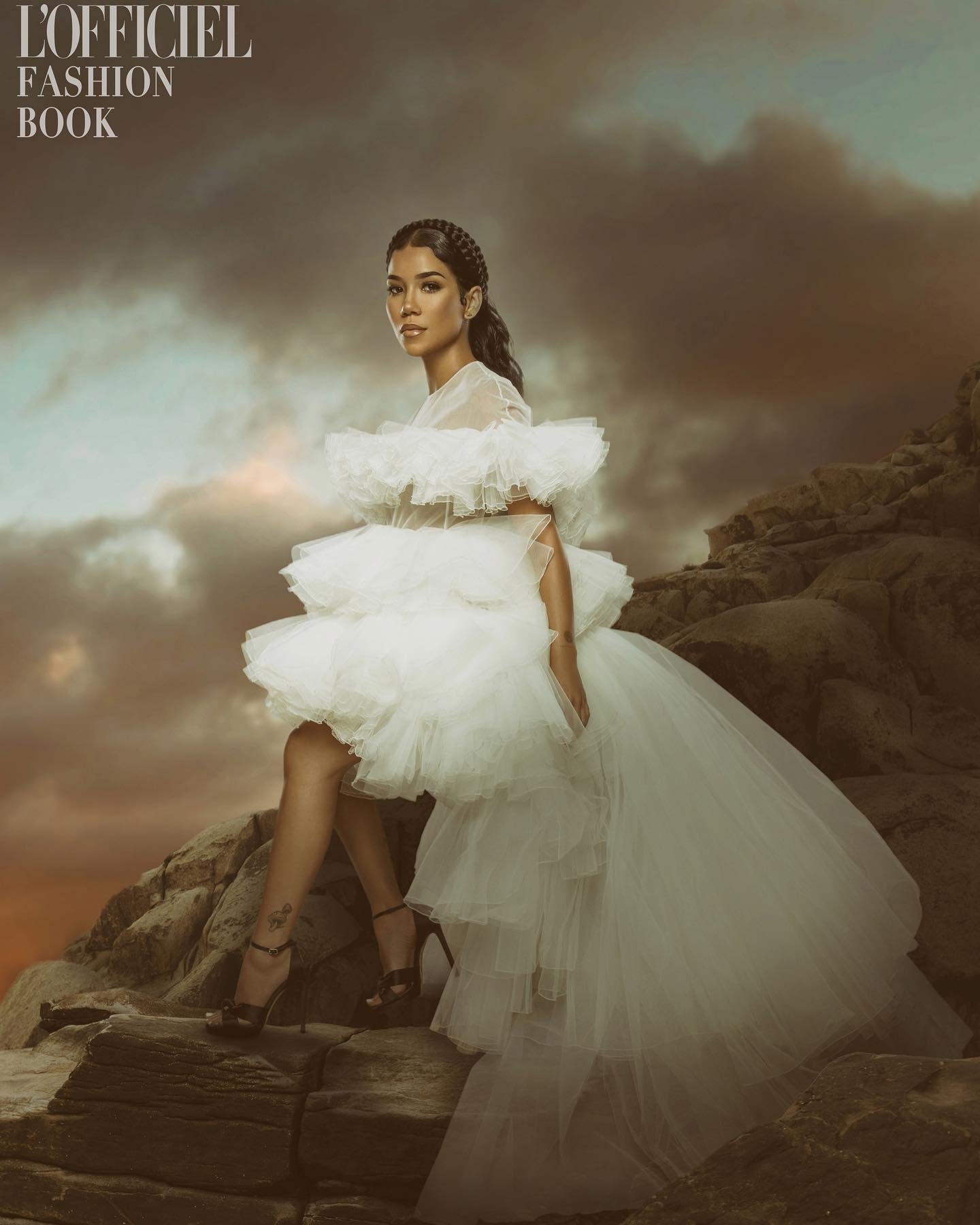 A woman in an elegant white gown poses on rocks against a dramatic sky in a fashion editorial for L'Officiel Fashion Book.