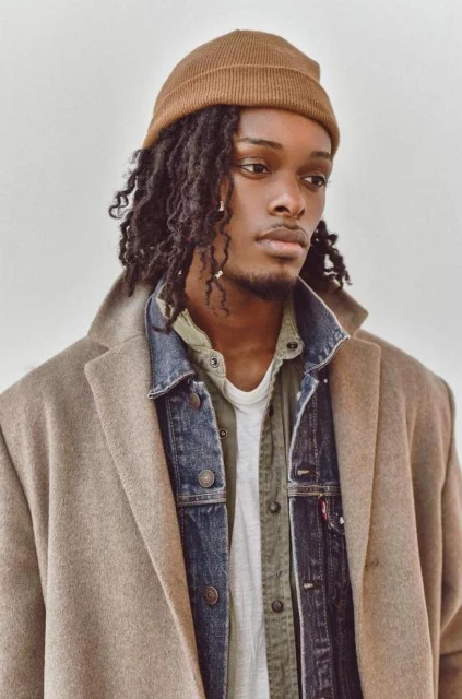 Portrait of a Black man with dreadlocks wearing a beanie, denim jacket, and coat against a white background in a studio setting.