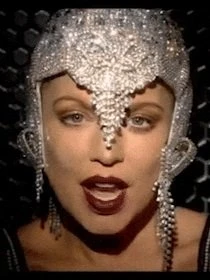 Close-up of an adult female with dark lipstick wearing an ornate, jeweled headdress against a dark, patterned backdrop.