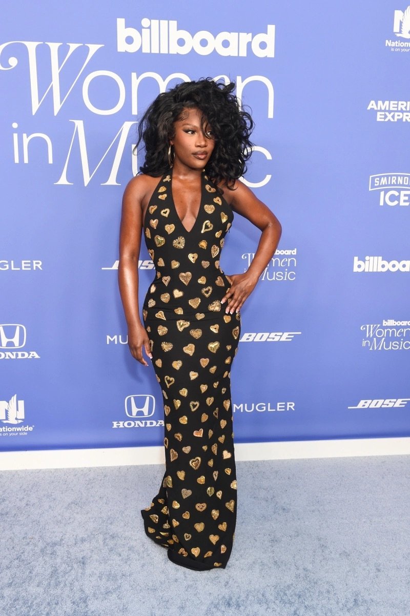 Black woman with voluminous dark hair in a black halter gown with gold hearts, standing on a carpet at an indoor event, looking at the camera.