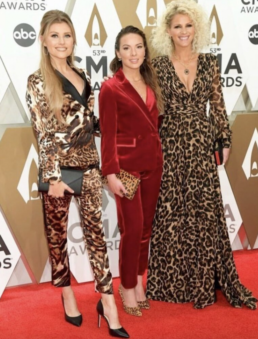 Three women pose on a red carpet at the 53rd CMA Awards. Two are wearing leopard print outfits and one is in a red velvet suit.