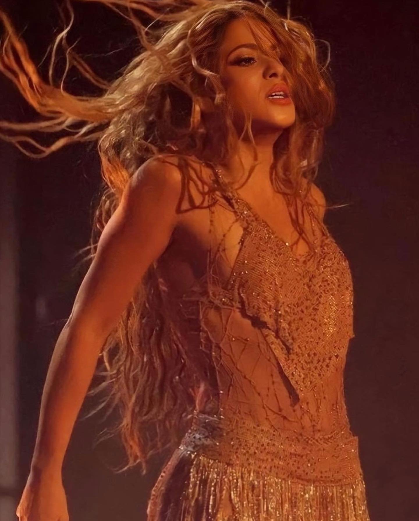 A woman with flowing blonde hair dances in a sparkly, fringed dress, captured in a medium shot with warm, stage-like lighting.
