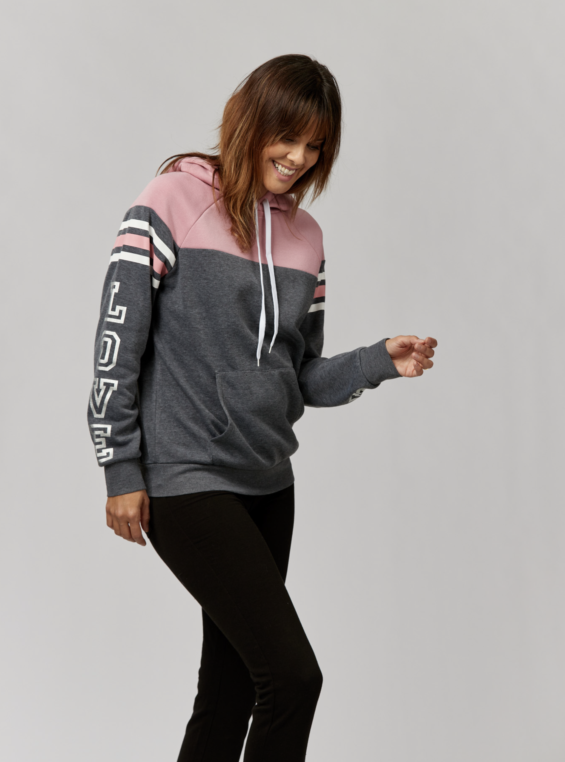 A smiling woman in a pink and gray hoodie stands in a neutral studio, showcasing a casual, comfortable style. Black leggings complete her look.