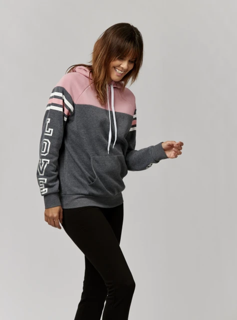 A smiling woman in a pink and gray hoodie stands in a neutral studio, showcasing a casual, comfortable style. Black leggings complete her look.