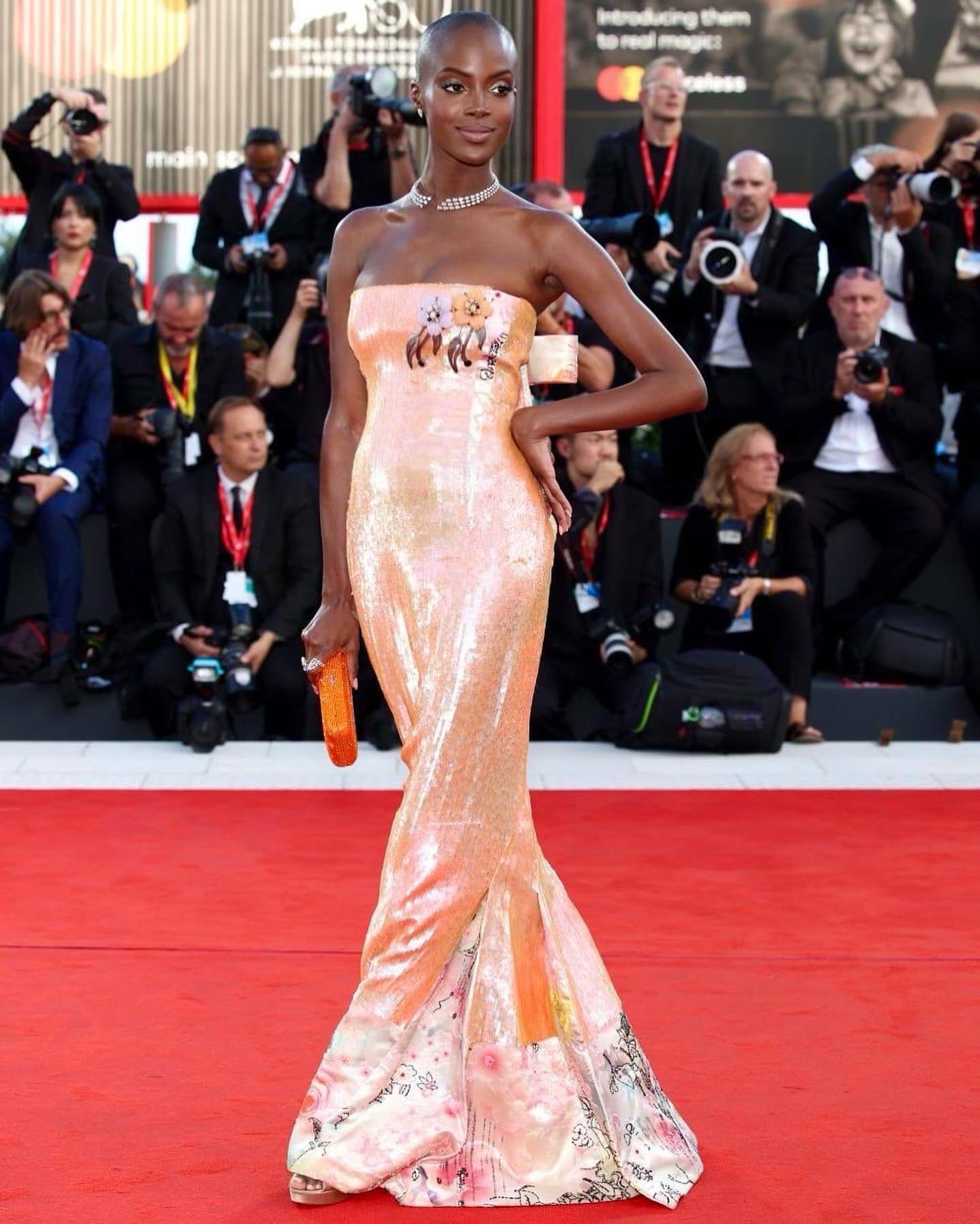 Woman in sequin gown with floral hem on red carpet at film festival, surrounded by photographers. Event style and celebrity fashion.