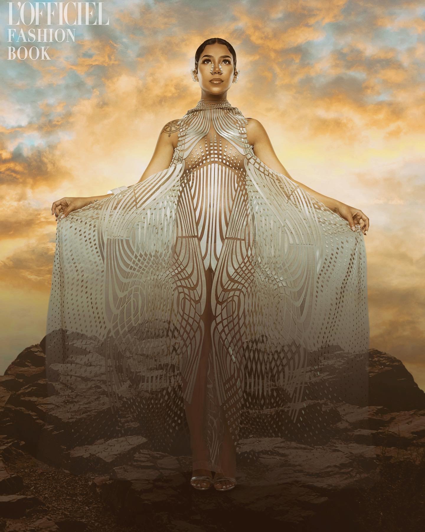 Fashion editorial portrait of a woman in an ornate, flowing gown posing against a dreamy sky and rocky landscape for L'Officiel Fashion Book.