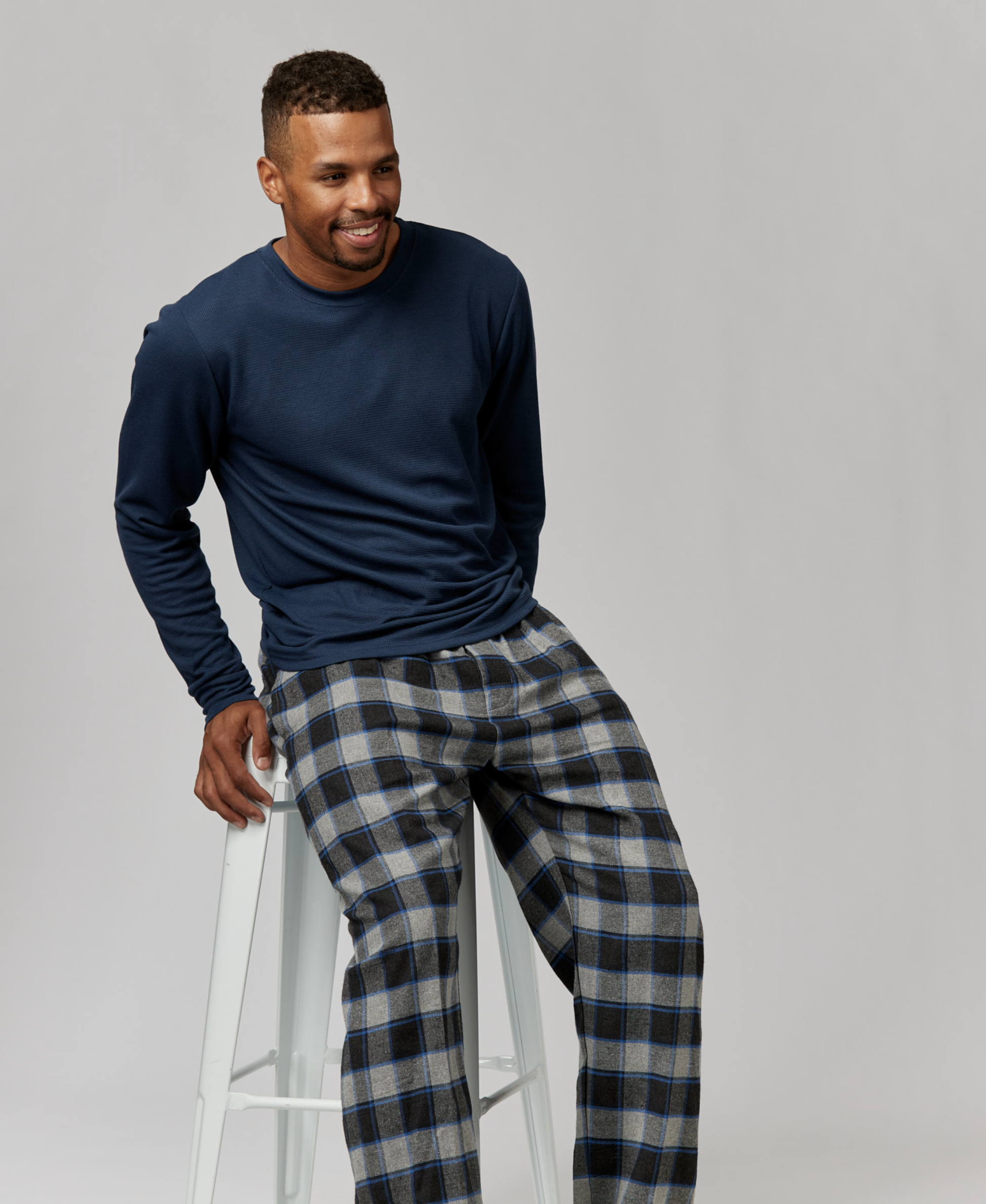 Man in a long-sleeved shirt and plaid pajama pants sitting on a white stool against a neutral backdrop in a studio setting.