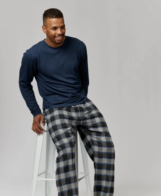 Man in a long-sleeved shirt and plaid pajama pants sitting on a white stool against a neutral backdrop in a studio setting.