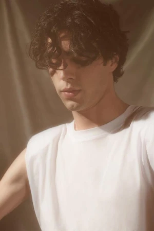 Close-up portrait of a young man with dark curly hair wearing a white t-shirt against a textured fabric background