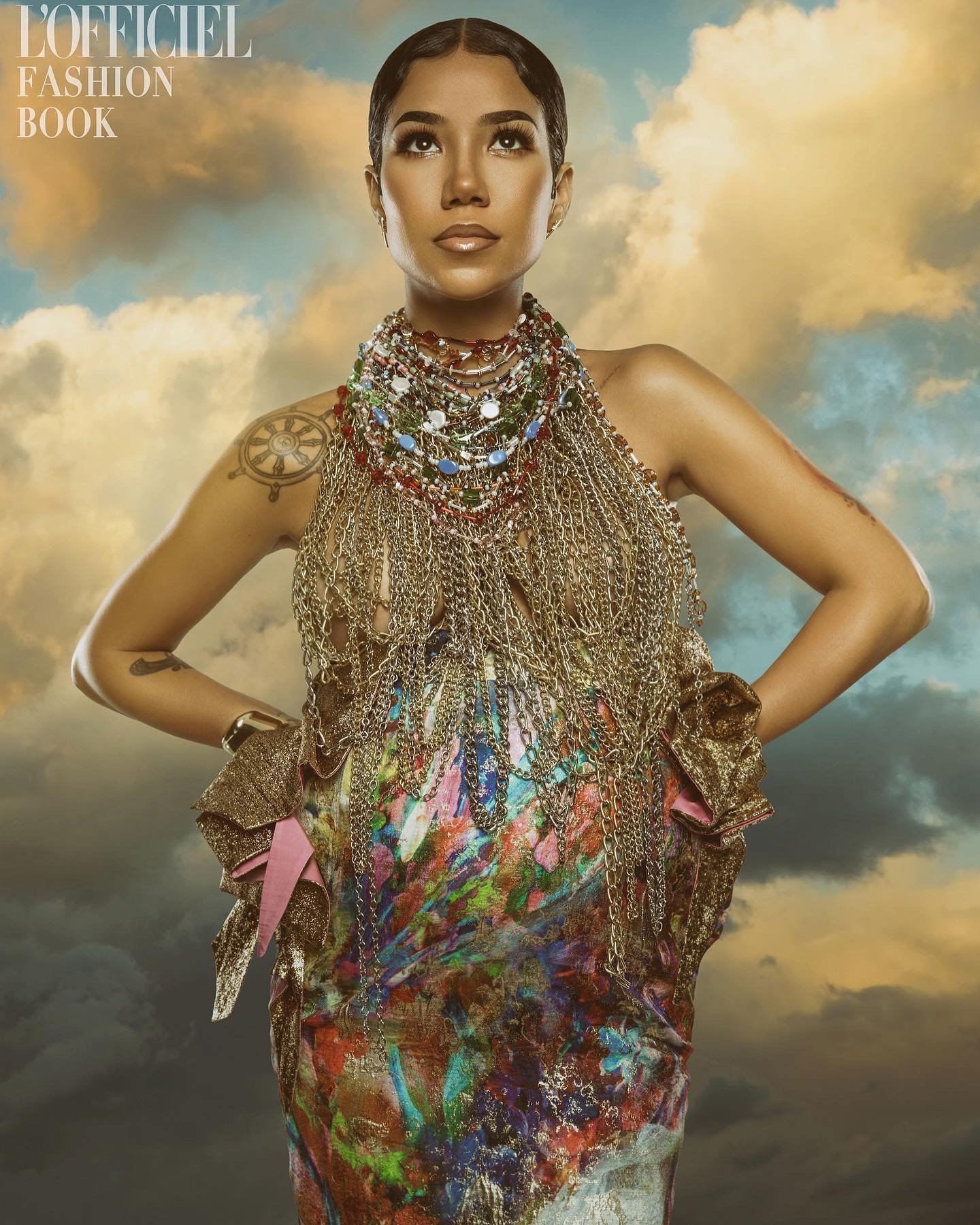 Jhene Aiko poses in a studio shot, wearing a gold chain top and colorful dress against a cloudy backdrop for a fashion magazine cover.