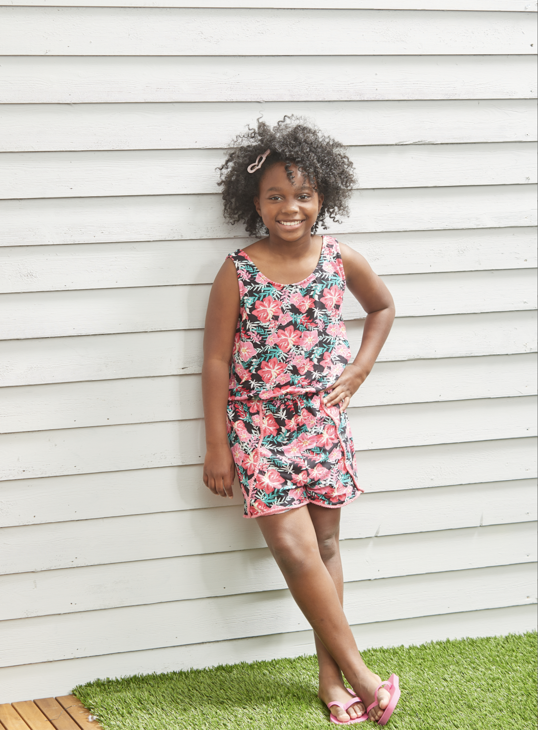 Portrait of a young girl wearing a floral dress. The girl stands against a white wooden fence in a backyard setting, smiling cheerfully.
