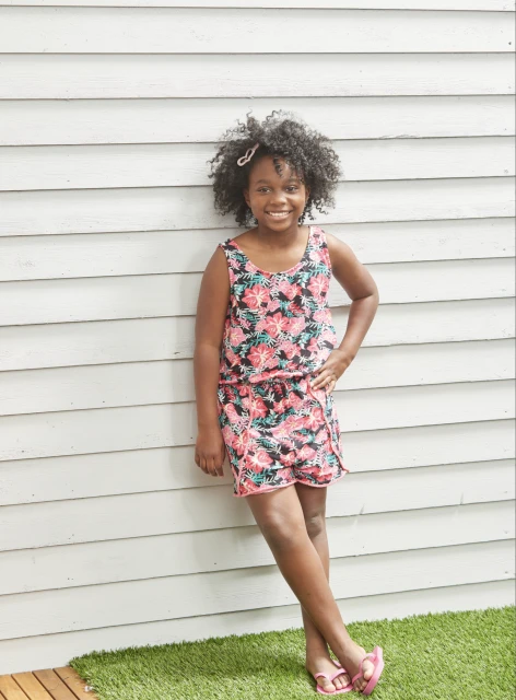 Portrait of a young girl wearing a floral dress. The girl stands against a white wooden fence in a backyard setting, smiling cheerfully.