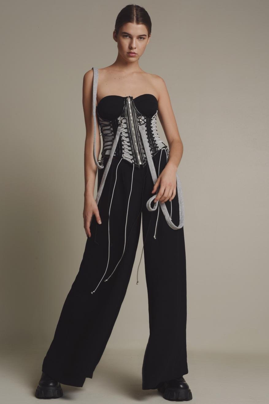 A woman poses in a strapless corset top and black trousers with white accents in a studio lit fashion shot, focusing on the clothing.