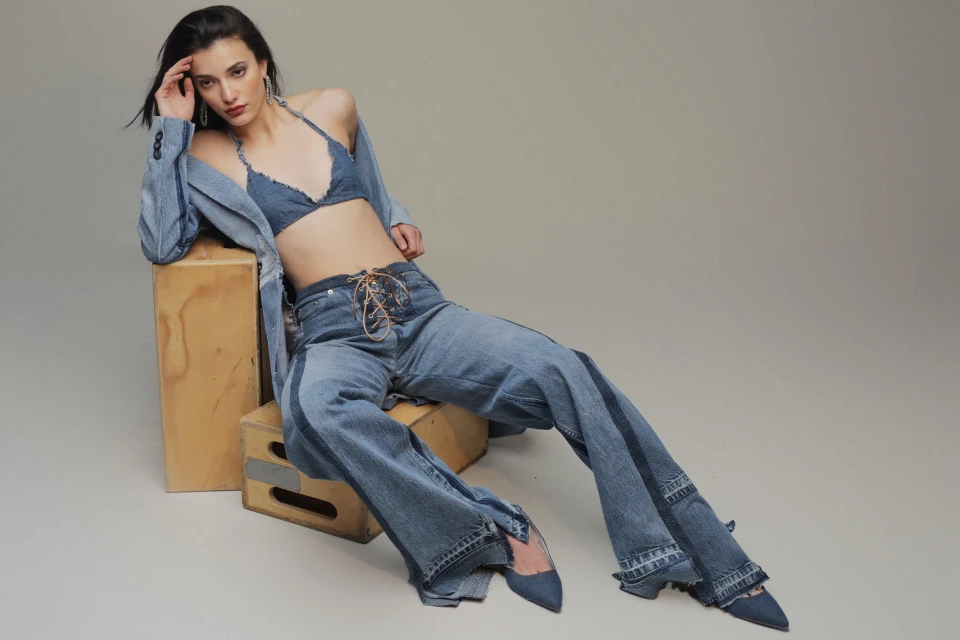 A young woman poses in a denim bra top, blazer, and wide-leg jeans, seated on wooden crates in a fashion editorial studio shot.