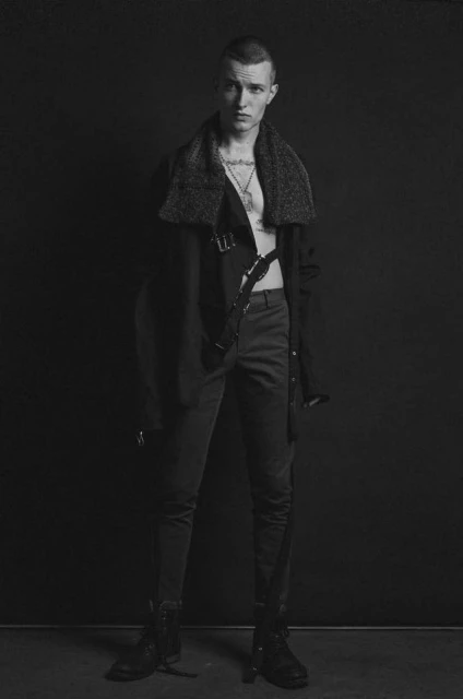 Monochrome portrait of a young adult male wearing a coat with a fur collar, pants, and boots, standing against a dark grey background.