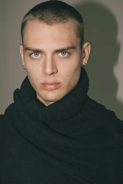 Close-up portrait of a young adult male with a buzzcut, wearing a black turtleneck sweater, set against a neutral background with soft lighting.