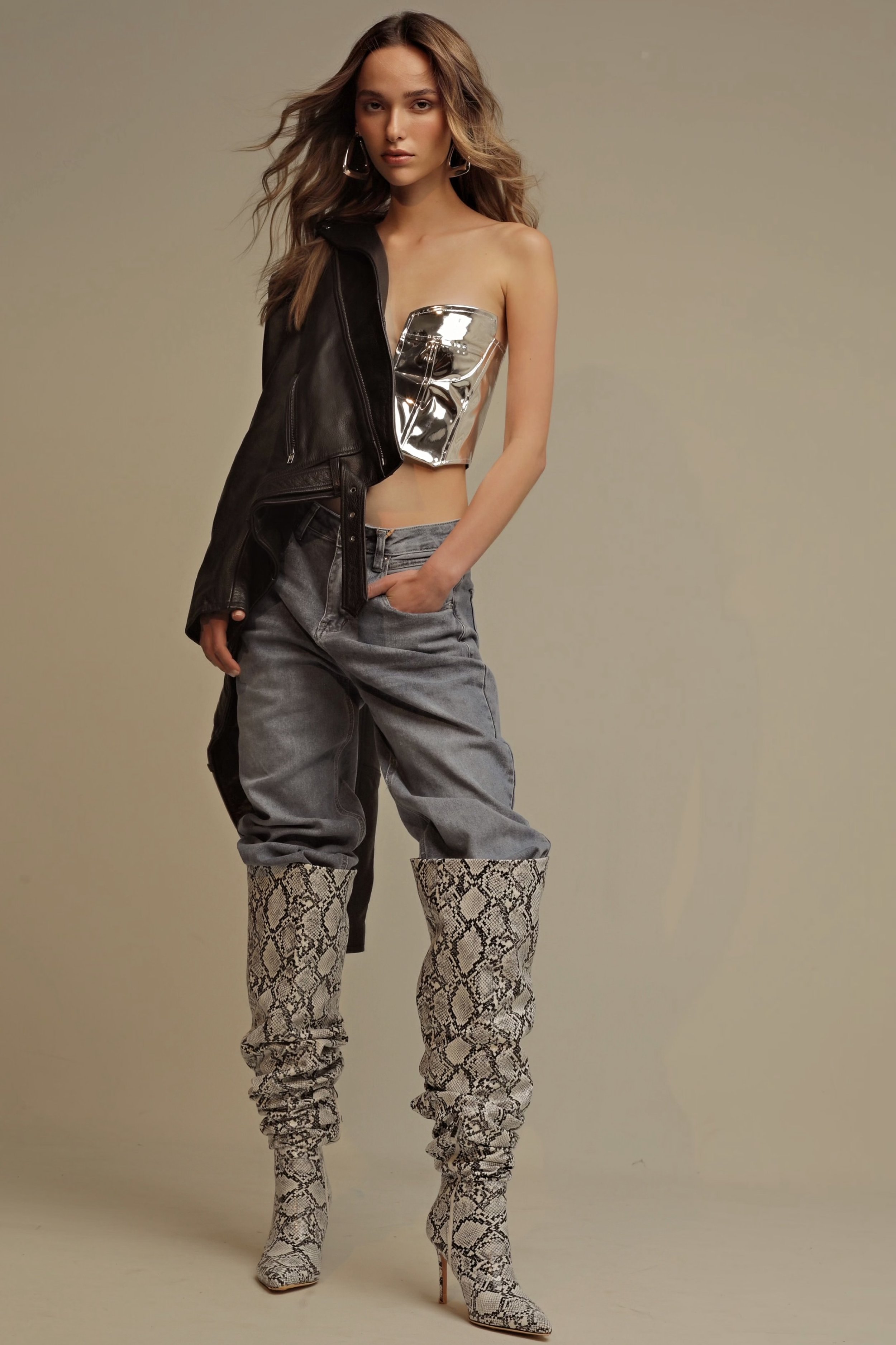 A woman in a futuristic silver corset, light blue jeans, and snakeskin boots stands in a studio, a black leather jacket on her shoulder.