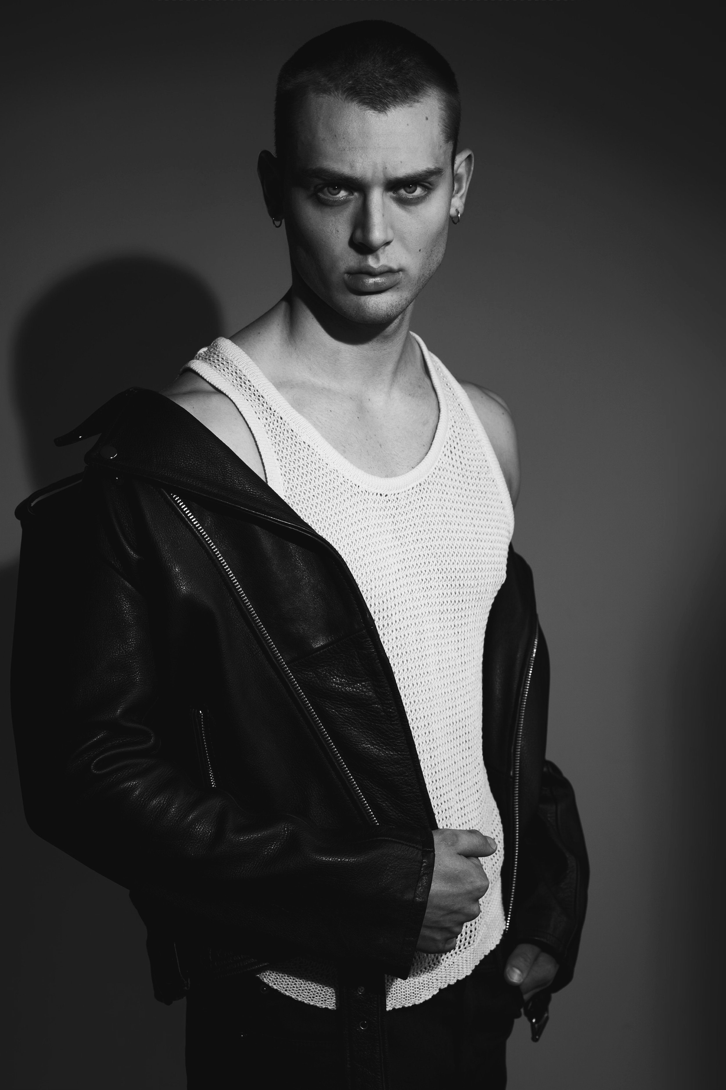 Black-and-white portrait of a young adult male with a shaved head, wearing a white tank top and a black leather jacket draped over his shoulders.