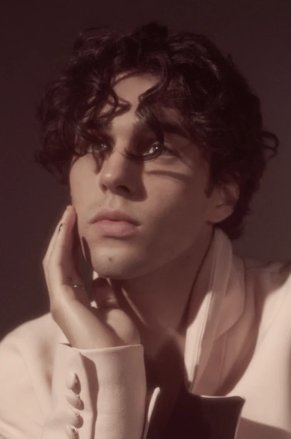 Close-up portrait of a young adult male with dark curly hair wearing a light pink jacket, striking a thoughtful pose with his hand.