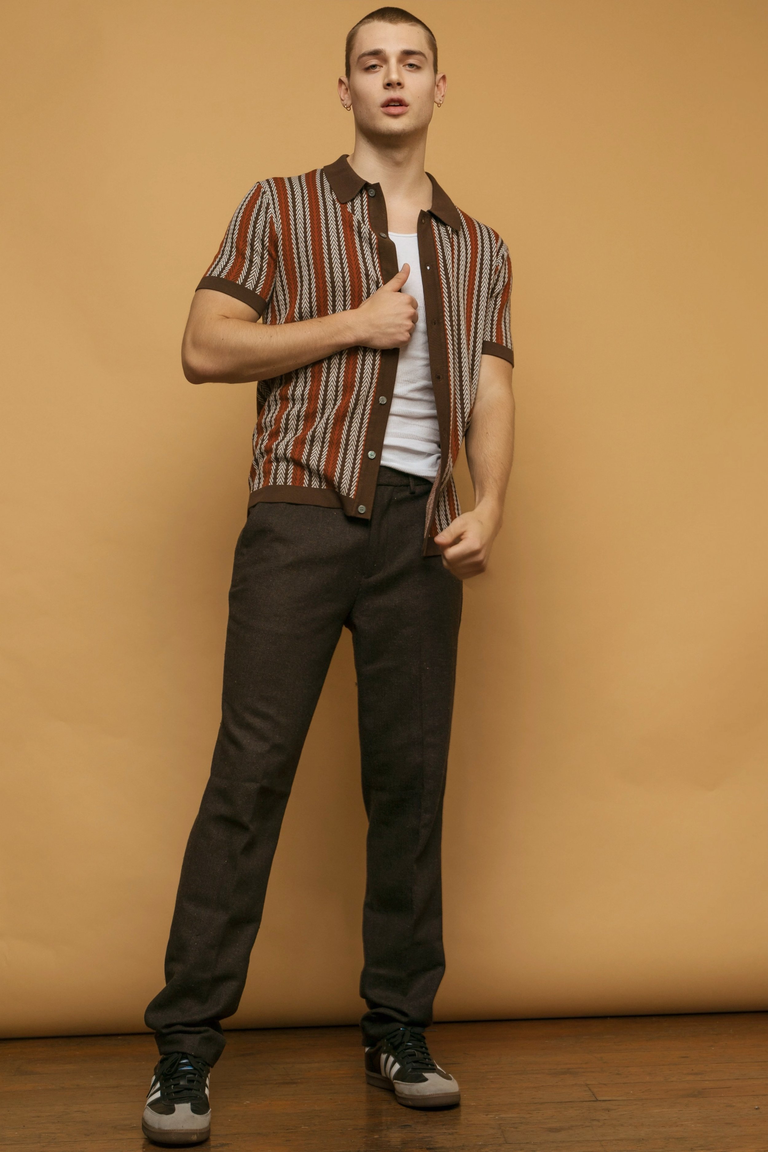 Full-length shot of a young adult male model wearing a striped collared shirt, dark pants, and sneakers, posing against a tan background.