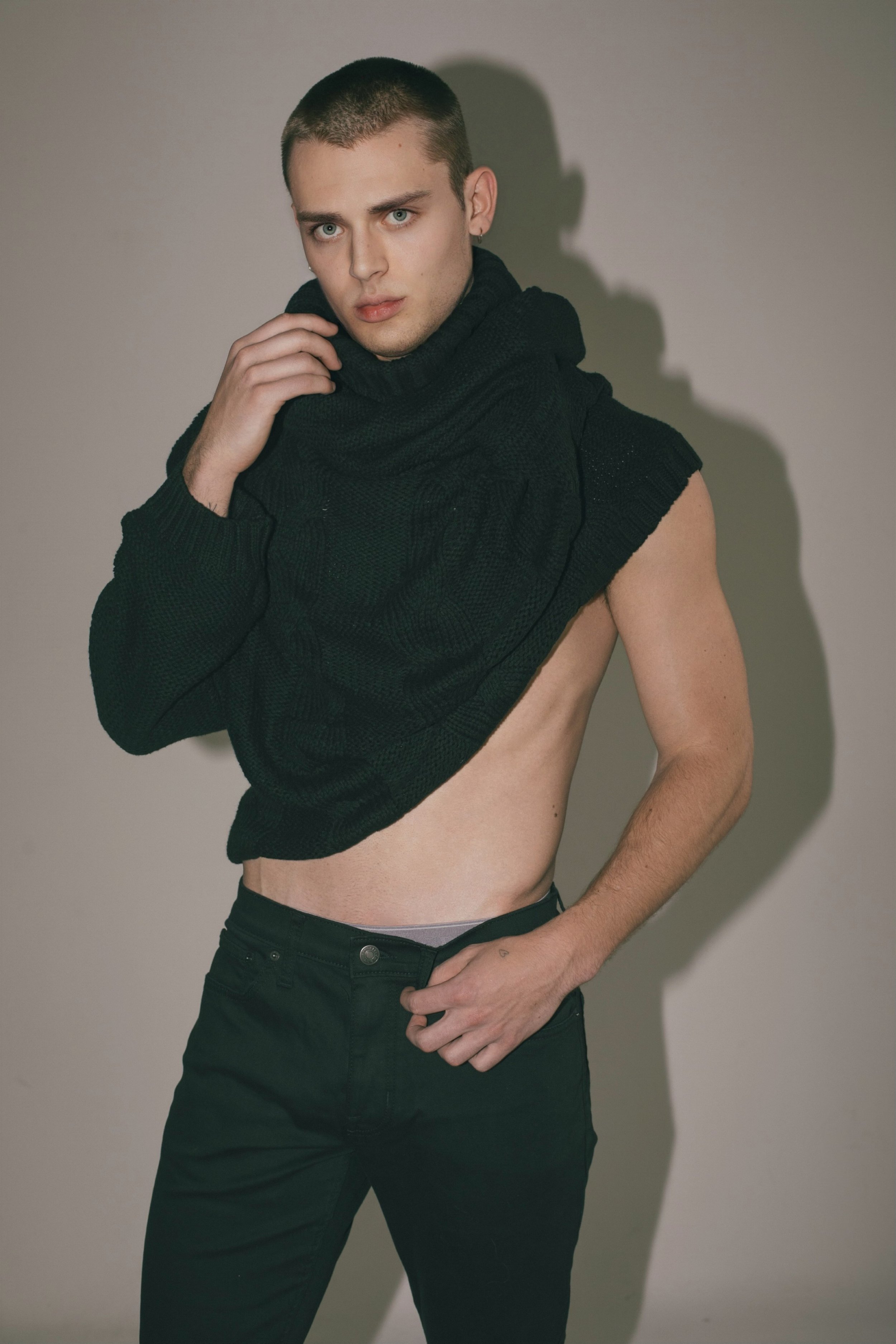 Young adult male posing in black jeans and a draped black turtleneck sweater in a studio setting, looking at the camera.