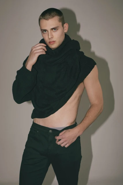 Young adult male posing in black jeans and a draped black turtleneck sweater in a studio setting, looking at the camera.