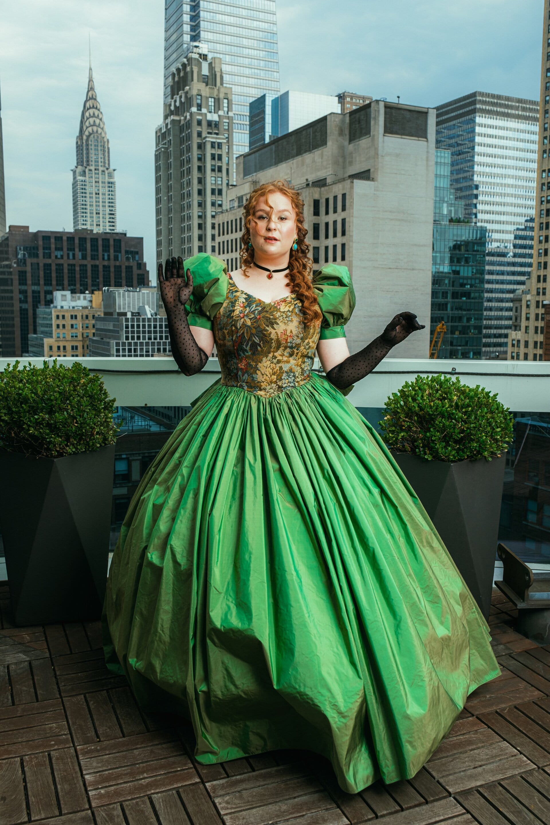 Woman poses in a green gown with puffy sleeves on a rooftop with a city skyline in the background on an overcast day.