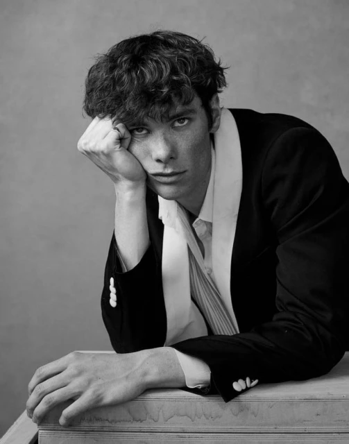 Monochrome portrait of a young adult male in a suit, resting his head on his hand, against a plain grey backdrop.