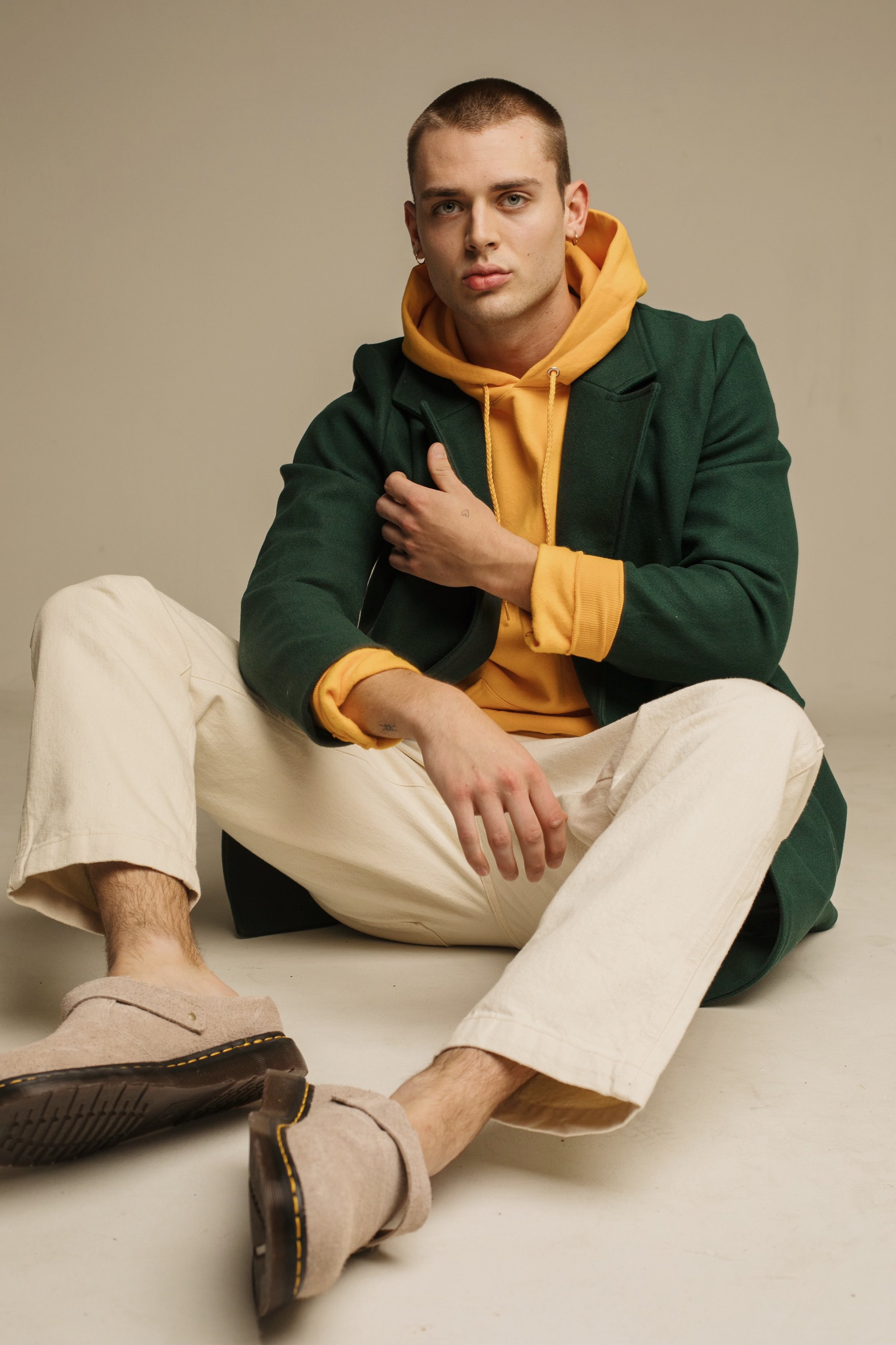 A young adult man in a yellow hoodie and green coat sits on the floor wearing casual cream-colored pants and beige Dr. Martens.