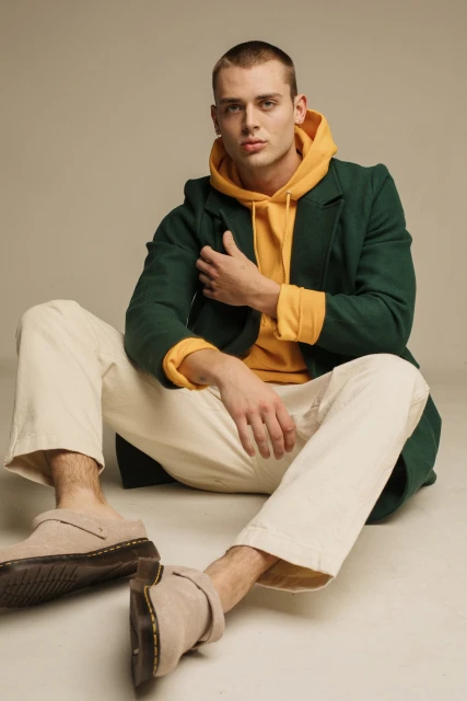 A young adult man in a yellow hoodie and green coat sits on the floor wearing casual cream-colored pants and beige Dr. Martens.